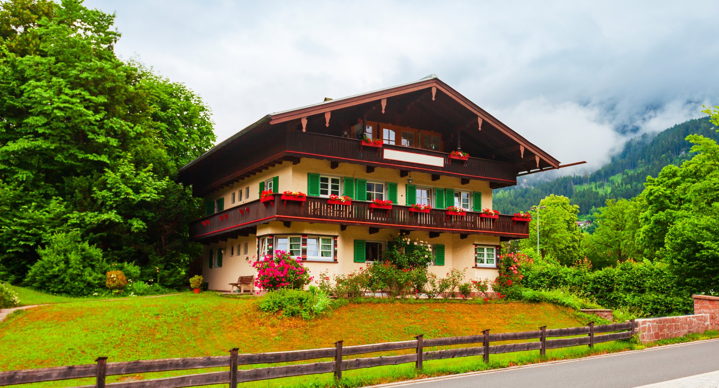 Oberbayerisches Wohnhaus mit Blumenbalkonen und Wiese vor bewaldetem Hang